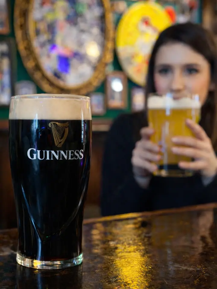 Una pinta de Guinness en primer plano y una clienta bebiendo una rubia detrás.