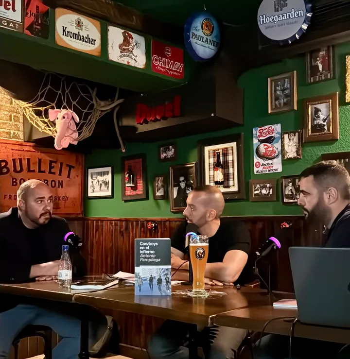 Tres hombres conversan ante micrófonos con una pinta de trigo y el libro «Cowboys en el infierno» sobre la mesa