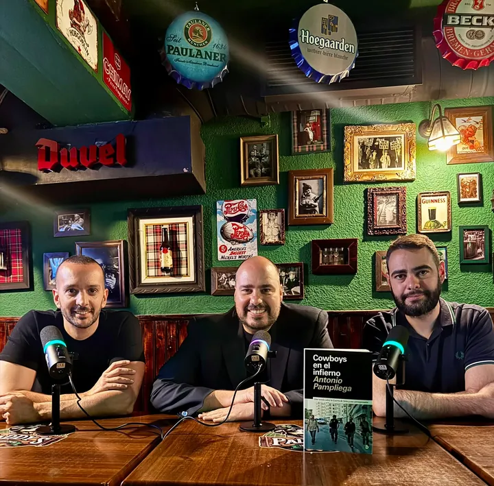 Tres hombres sonríen tras tres micrófonos en una mesa del bar; delante, el libro «Cowboys en el infierno» de Antonio Pampliega y la pared verde llena de cuadros