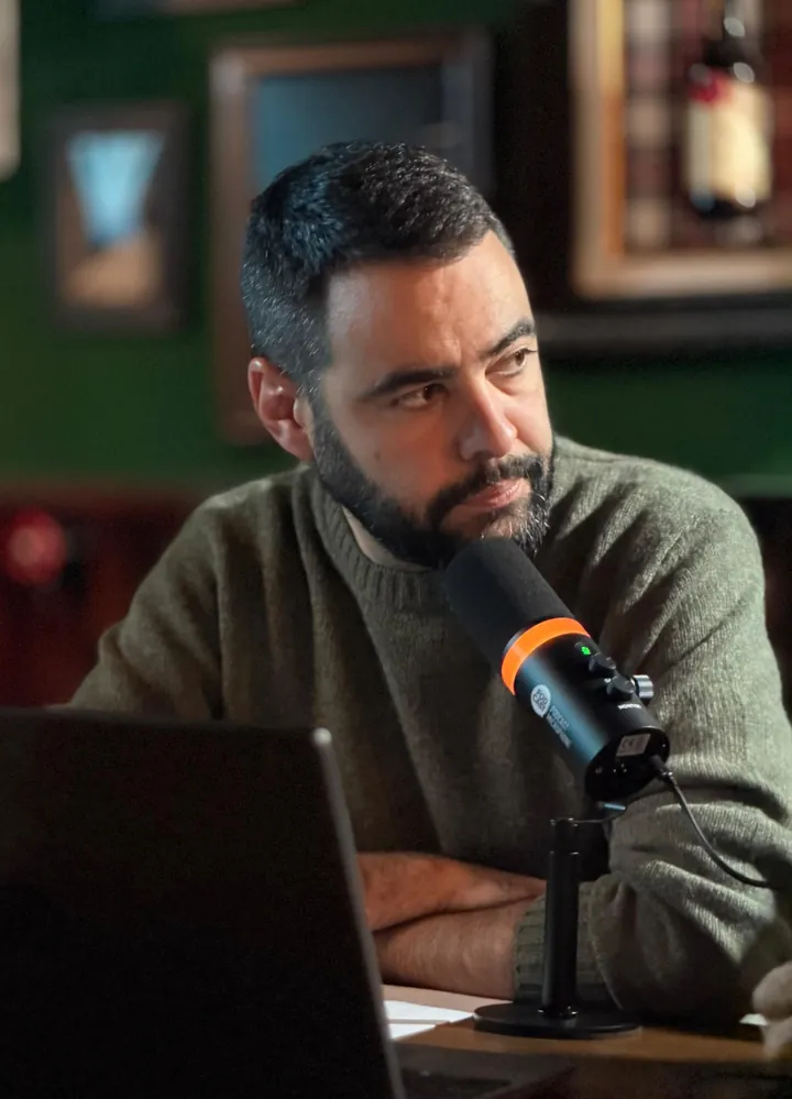 Un presentador con barba y jersey verde escucha con los brazos cruzados ante un micrófono