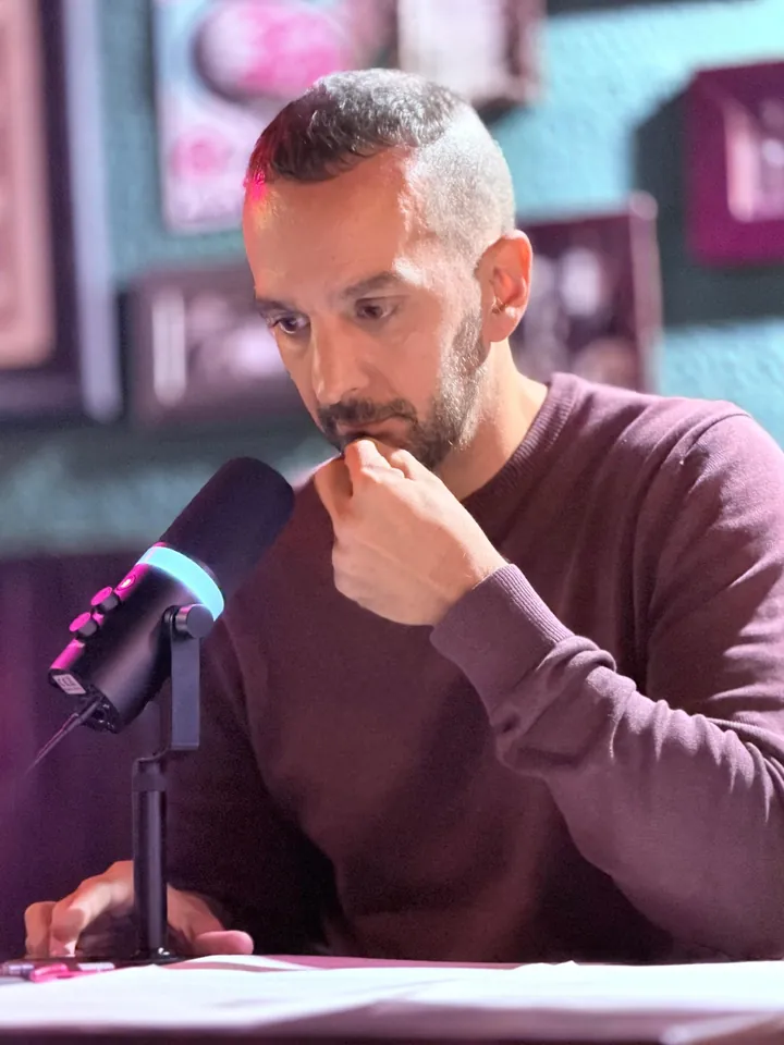 Un presentador con jersey granate repasa sus notas con la mano en la barbilla, junto al micrófono