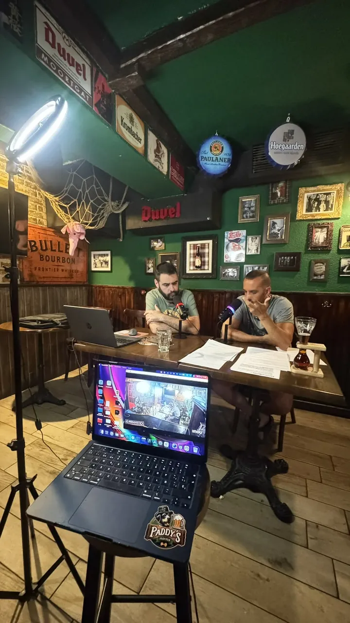 Dos presentadores graban en una mesa del bar; en primer plano, un foco de aro y un portátil con la pegatina de Paddy’s